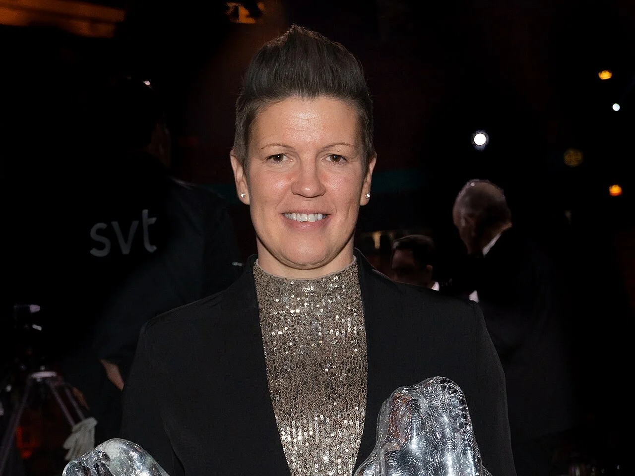 Erika Holst ler i svart kavaj och paljettröja med två glaspriser i händerna, fotograferad 2022